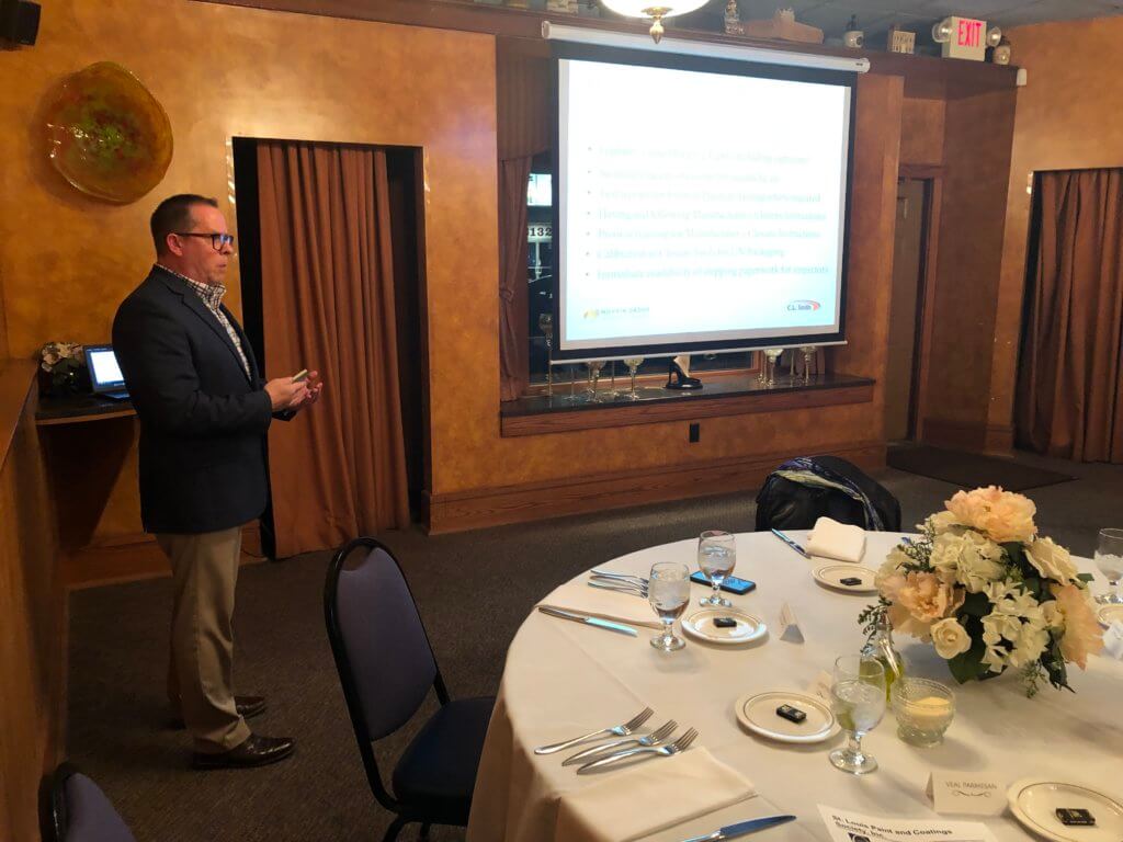 A person from the St. Louis Paint & Coating Society stands next to a presentation screen in a dimly lit room, giving a talk to an unseen audience. A table in the foreground is set for a meal, with a flower centerpiece, plates, cutlery, and glasses.