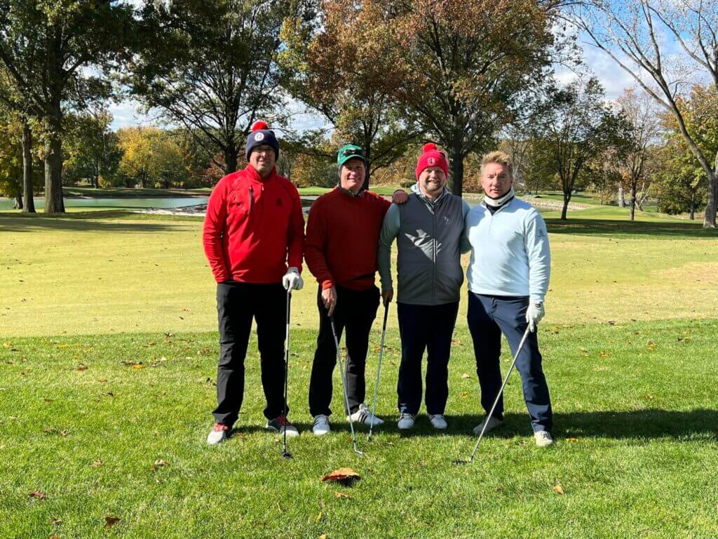 Four men stand on a golf course, each holding a golf club, participating in the SLPCS Fall Golf Tournament 2023. They're dressed in jackets and hats, suggesting cool weather. Trees with autumn leaves surround the grassy area, and a sand trap is visible in the background.