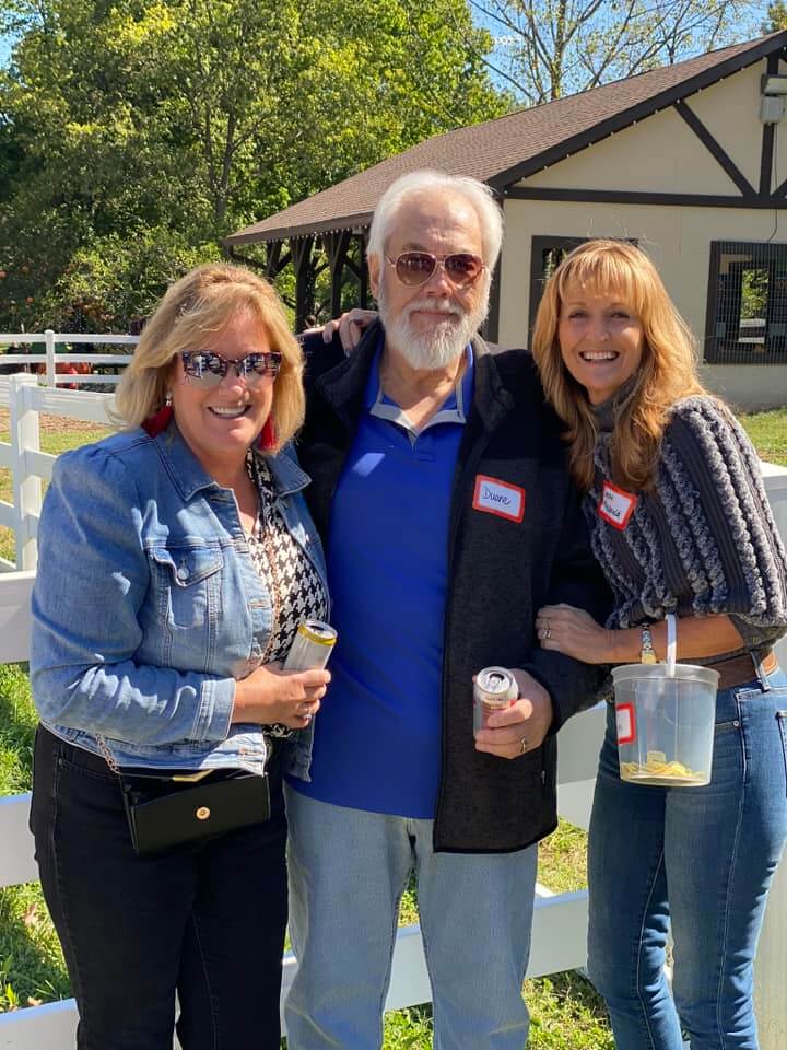 Three people stand outside near a white fence and a rustic building at the Company Picnic 2023. The man in the center wears sunglasses and a name tag, holding a can. The women beside him are smiling, one with a drink, the other with a jar. It's a sunny day with trees in the background.