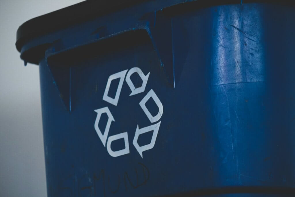 Close-up of a blue recycling bin with a white recycling symbol on its side. The surface appears slightly worn, and there is faint writing below the symbol. The background is blurred.