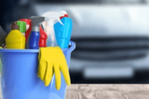 A blue bucket filled with various cleaning spray bottles and a yellow glove sits on a wooden surface. The background shows a blurred car front.