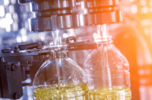Close-up of a bottling machine filling clear plastic bottles with a yellow liquid, possibly oil or juice. The machinery is metallic and in focus, with a warm orange glow in the background, suggesting an industrial setting.