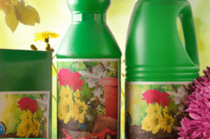 Close-up of gardening supplies with vibrant floral labels. The image features green containers displaying colorful flower images, surrounded by pink and yellow blooms. In the background, gardening tools and a leaf are visible.