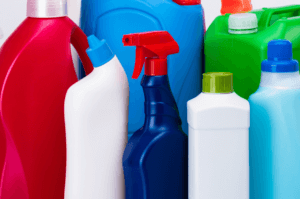 Various colorful plastic bottles and containers for household cleaning products, including red, blue, green, and white bottles. Some have spray nozzles, while others have screw caps. The arrangement is close together.