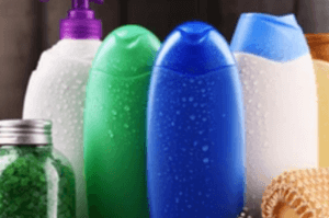 A collection of colorful shampoo and conditioner bottles, including green, blue, and white, stand upright. A jar with green bath salts and a brown loofah are placed beside them. The bottles have a light layer of condensation.