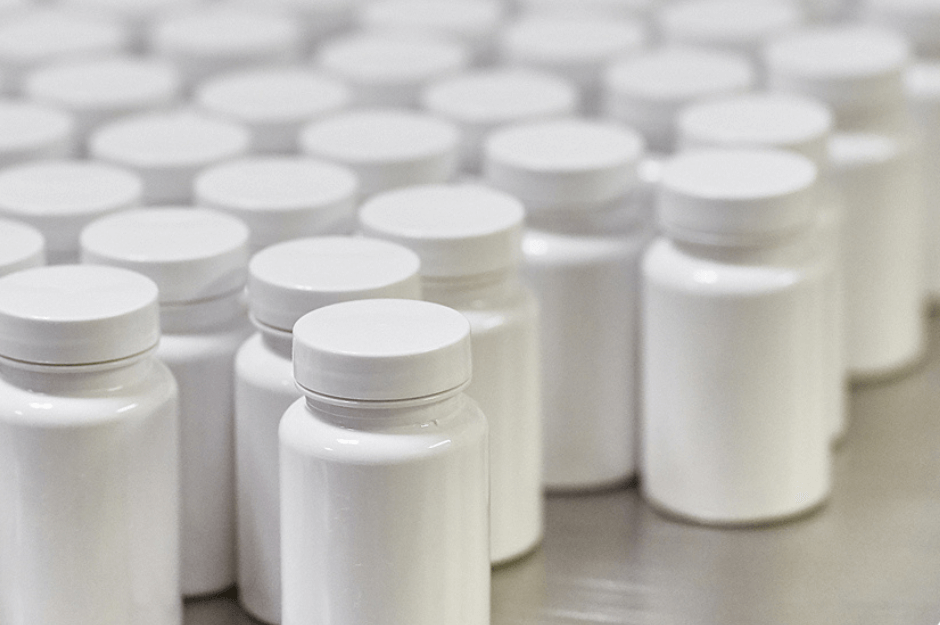 Rows of white plastic bottles with white caps arranged closely together on a flat surface. The bottles are uniform in size and shape, suggesting a mass production setting.