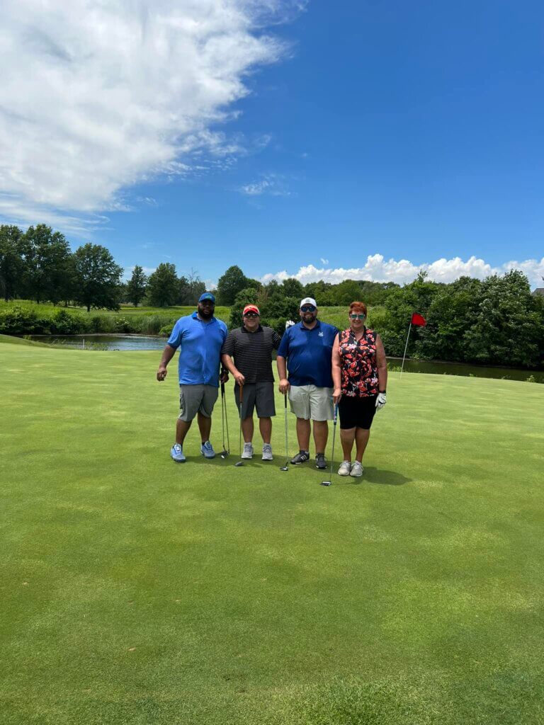 Four participants line up on the lush green of the golf course, clubs in hand, as part of the SLPCS Spring Golf Tournament 2024. Behind them, serene trees and a tranquil pond complement the blue sky dotted with clouds.
