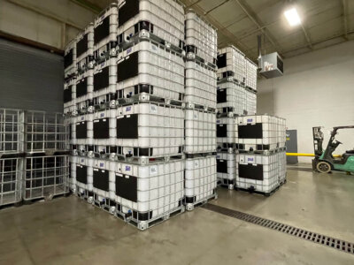 Stacked industrial containers in a warehouse, organized neatly in rows. A forklift is visible in the background, and the space has a large, open layout with a high ceiling and concrete floor.