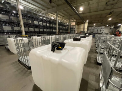 A large warehouse filled with neatly arranged white plastic containers secured in metal cages. The containers are lined up on the floor, with more stacked along the walls. The space is well-lit with industrial lighting.