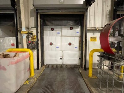 Loading bay with large white containers stacked in an open dock door marked "3". Yellow safety rails flank the area. A red industrial fan is on the right, and a large bin is on the left. The setting appears to be an industrial warehouse.