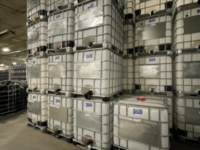 Stacks of large, square liquid containers with metal frames organized in a warehouse. The containers are labeled, and the floor and ceiling are concrete. The setting appears industrial and spacious.