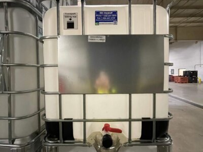 A large white industrial container with a metal cage and a red valve is stacked on top of another. It is labeled with a sign and set in a warehouse environment with other containers nearby.
