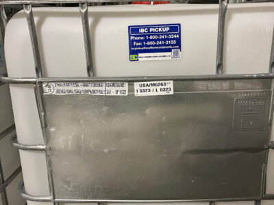 Stacked IBC containers, one prominently labeled with contact info. A barcode and detailed text are visible. Metal cage surrounds the white container, which has a plastic valve at the bottom.
