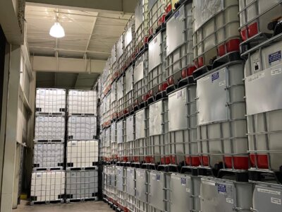 A warehouse aisle filled with industrial containers stacked in rows. The containers are large, square, and gray with red bases, secured in metal frames. The ceiling is high with visible lighting fixtures. The floor is smooth concrete.