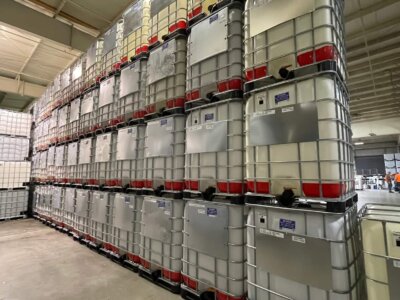 Rows of large, stacked industrial containers in a warehouse with high ceilings. The containers are gray with red tops and supported by metal frames. A concrete floor and shelves in the background are visible.