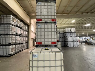 Tall stack of industrial containers, each with a metal grid frame, inside a large warehouse. The floor is concrete, and similar stacks are visible in the background, suggesting storage or distribution.