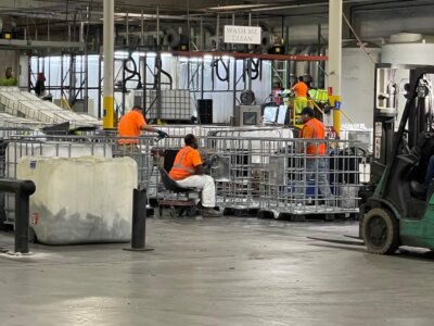 Workers in bright orange safety gear tend to equipment and containers in a large industrial warehouse. A forklift is nearby, and overhead fixtures provide lighting. The space is organized with metal structures and visible signage.