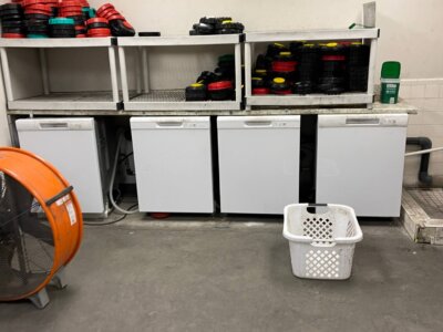 A storage room with three white mini-fridges on the floor. Above them, shelves hold stacks of red and black circular weights. A large orange industrial fan is on the left, and a white laundry basket is on the floor.