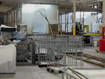 A large industrial warehouse with metal beams, plastic crates, pallets, and equipment scattered throughout. Bright overhead lighting illuminates the space. White and transparent plastic sheets hang in the background.