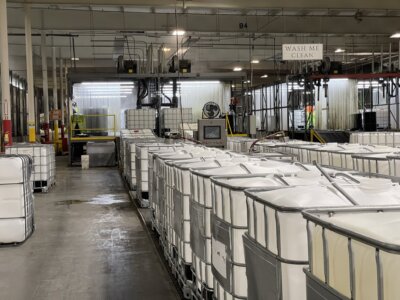 Rows of large white industrial containers are lined up in a factory setting. Behind them, machinery and a washing area with a sign saying "WASH ME CLEAN" are visible. The space is well-lit with overhead lighting.