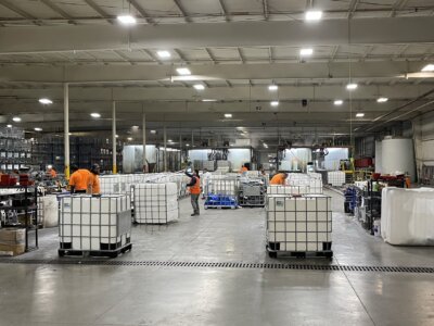 Large industrial warehouse interior with workers in orange shirts organizing large white storage containers. Shelves with various items line the walls, and bright overhead lights illuminate the space.