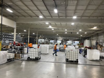 Warehouse interior with several workers in orange shirts moving among large, cube-shaped containers on pallets. Shelves line the walls, and fluorescent lights illuminate the high-ceilinged space.