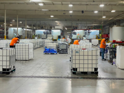 Workers in orange shirts are in a large industrial facility, handling large white containers on pallets. The spacious room is well-lit, with various equipment and shelving in the background. Signs and safety gear are visible throughout.