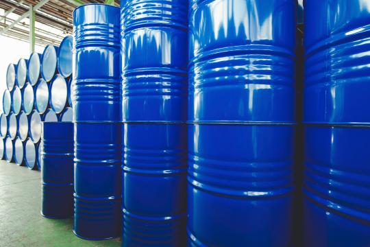Several rows of blue industrial barrels are stacked vertically in a warehouse. The lighting is bright, casting reflections on the shiny metal surfaces. The background shows more barrels and part of the warehouse's ceiling structure.