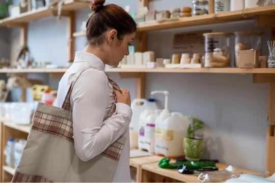 A person with a plaid tote bag browses a shelf of eco-friendly products, including jars and bottles, in a store. They are wearing a white shirt and have their hair tied back.