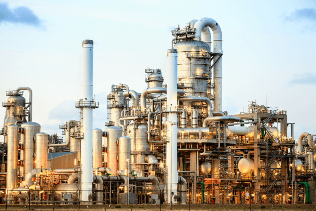 Industrial chemical plant with numerous interconnected pipes, towers, and storage tanks. The metallic structures glisten under daylight, showcasing a complex network of machinery against a clear sky.