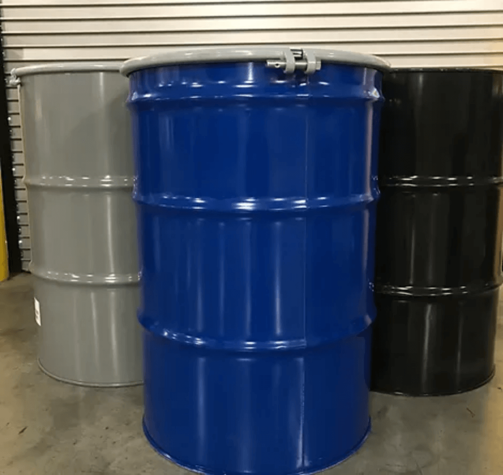 Three metal barrels stand in a row on a concrete floor. The barrels are colored gray, blue, and black from left to right. A metal pipe rests on top of the blue barrel. The background features a closed metal shutter.
