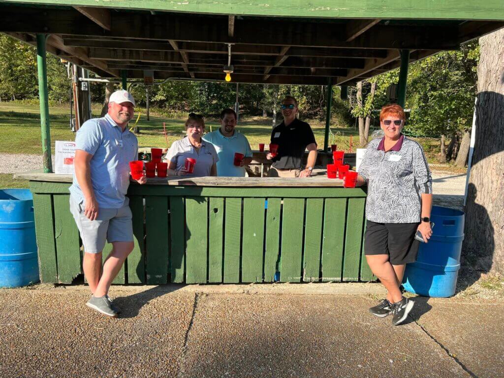 Five people stand around a green wooden counter outdoors, holding red cups at the SLPCS Annual BBQ. They are under a rustic shelter with trees and grass in the background, surrounded by blue barrels. It's a sunny day, perfect for celebrating togetherness and good company.