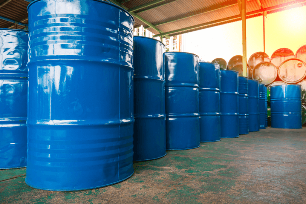 Rows of blue industrial barrels are neatly arranged in a warehouse with a corrugated metal roof. Sunlight streams in from the right, illuminating the metallic surface of the barrels and casting shadows on the concrete floor.