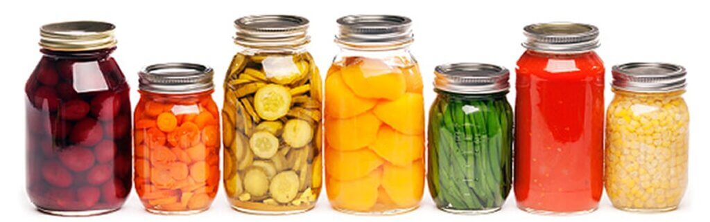 Seven jars of preserved produce arranged in a row, featuring different contents: red beets, orange carrots, green pickles, sliced peaches, green beans, red tomatoes, and yellow corn. Each jar has a metal lid.