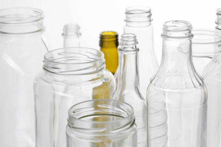 A variety of empty glass jars and bottles in different shapes and sizes are clustered together against a white background. They are mostly clear, with one brown bottle among them.