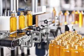 An industrial bottling line is filling transparent bottles with yellow liquid. The bottles, capped with golden lids, are moving along the conveyor belt amid machinery, with many filled bottles visible in the foreground.