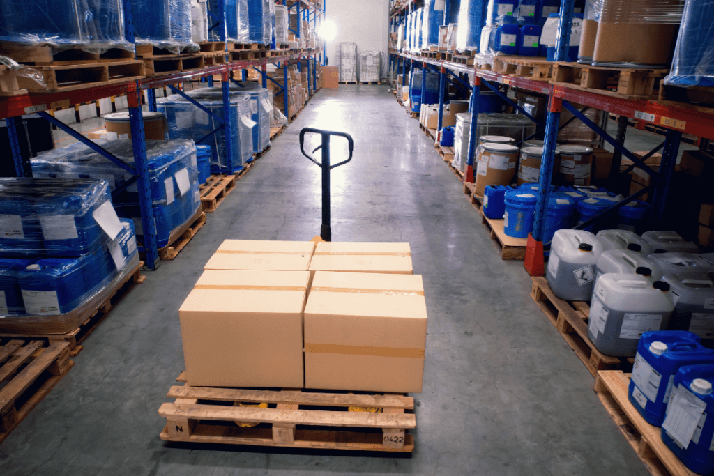 A pallet jack holds cardboard boxes on a wooden pallet in the center aisle of a warehouse. Shelves filled with blue and white containers line both sides. The warehouse has a polished concrete floor and high ceiling.