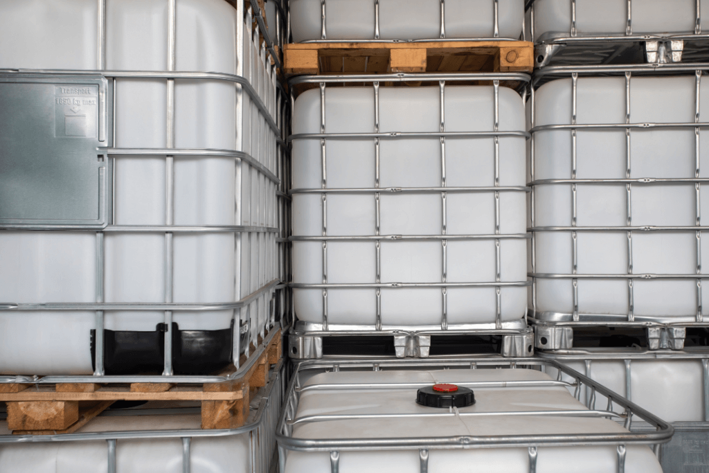 Stacks of large, industrial plastic containers with metal frames are shown on wooden pallets, arranged in a warehouse. The containers are white with a visible valve on the top of one.
