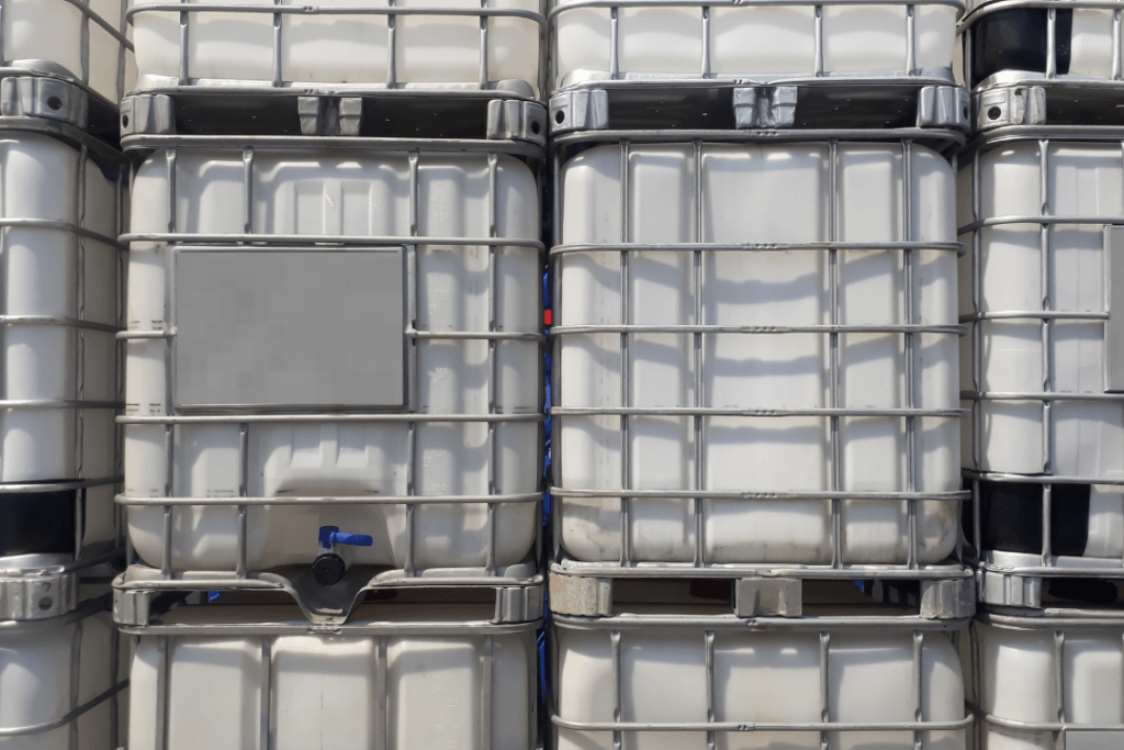 Stacked industrial containers with a grid-like metal frame, used for liquid storage. Each container has a spout at the bottom. The containers are uniform, lined up closely together, and shaded in white and gray tones.