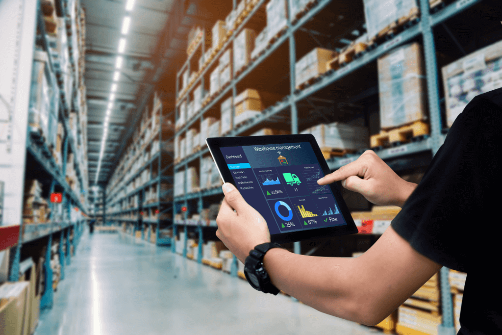 A person using a tablet in a large warehouse filled with shelves stocked with boxes. The tablet displays graphs and data analytics related to inventory management. The scene conveys a sense of modern logistics and technology.