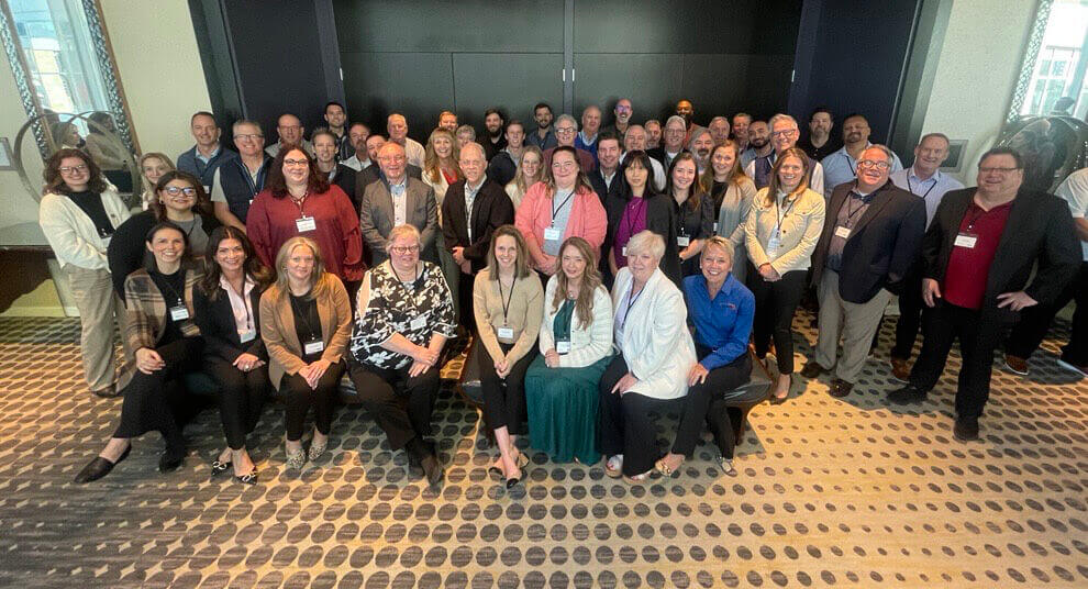 A large diverse group of professionally dressed people is posing at the Novvia Group Annual Leadership & Sales Conference. They are smiling and standing on a patterned carpet, facing the camera, with dark panel doors in the background.