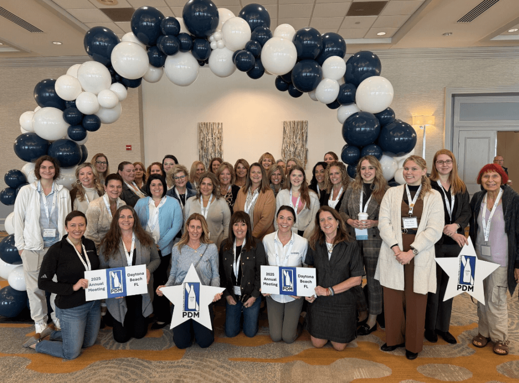 A group of smiling people pose under a blue and white balloon arch indoors. Some are holding signs that read 