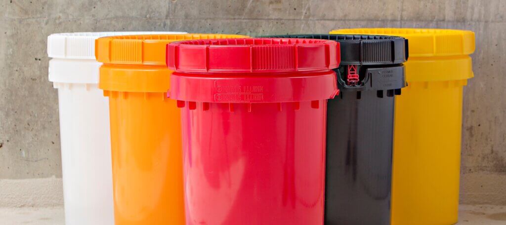Four large plastic barrels with lids in white, orange, red, and black are positioned in a row on a concrete floor against a concrete wall. The barrels are clean and appear unused.