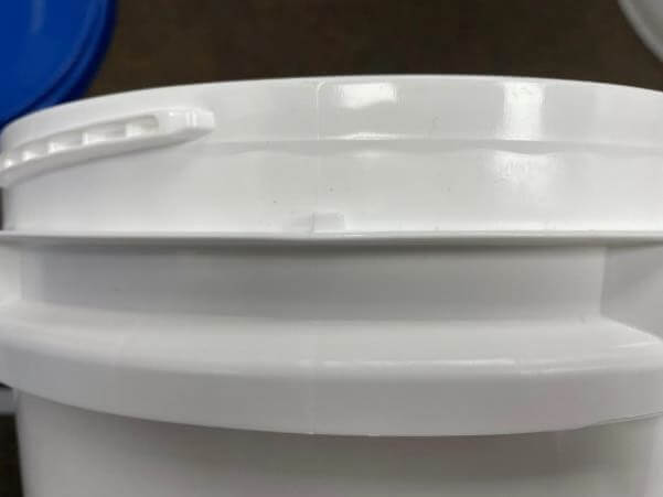 Close-up of the top section of a white plastic bucket with a handle. The surface appears smooth and shiny under the lighting. Part of a blue lid is visible in the background.