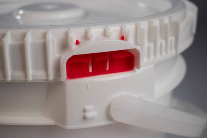 Close-up of a white plastic container lid with a red locking mechanism. The lid features multiple grooves and a small handle on the side. The background is blurred.