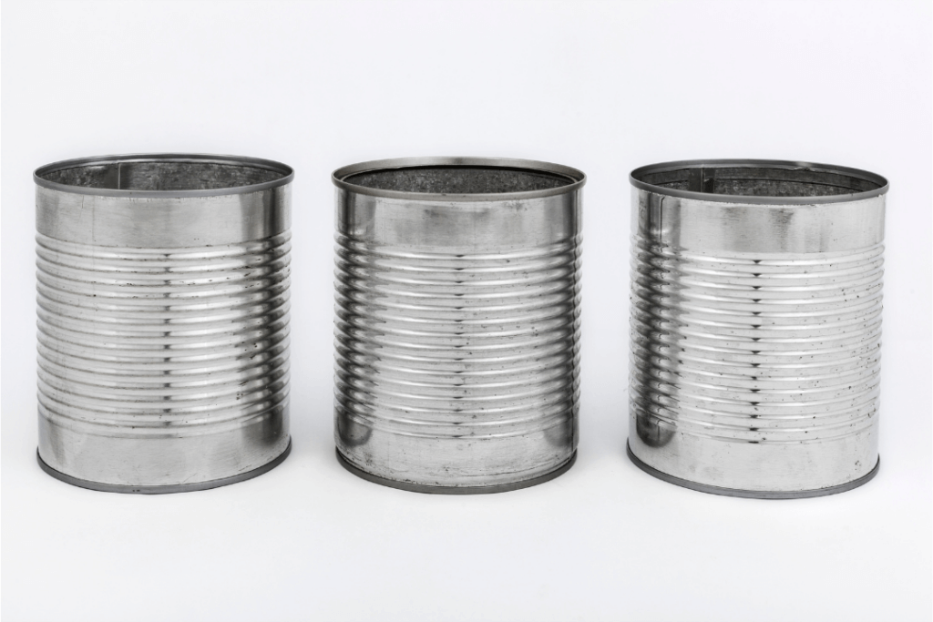Three empty, silver tin cans with ridged surfaces are aligned side by side against a plain white background.