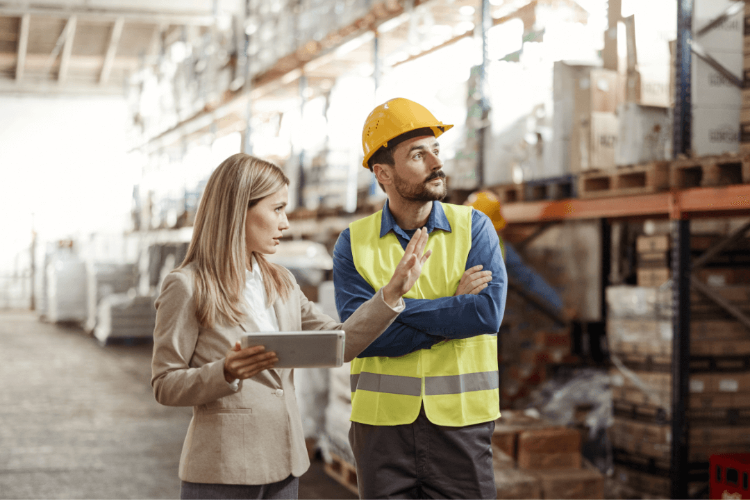 A woman holding a tablet talks to a man in a hard hat and safety vest inside a warehouse. They are surrounded by shelves stacked with boxes and supplies.