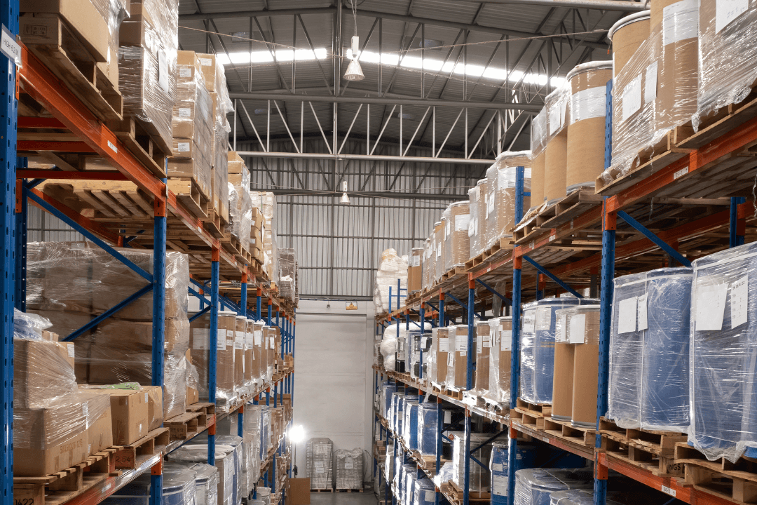 Rows of tall warehouse shelves filled with pallets of wrapped goods and containers, under a high metal ceiling with fluorescent lights, creating an organized industrial storage environment.