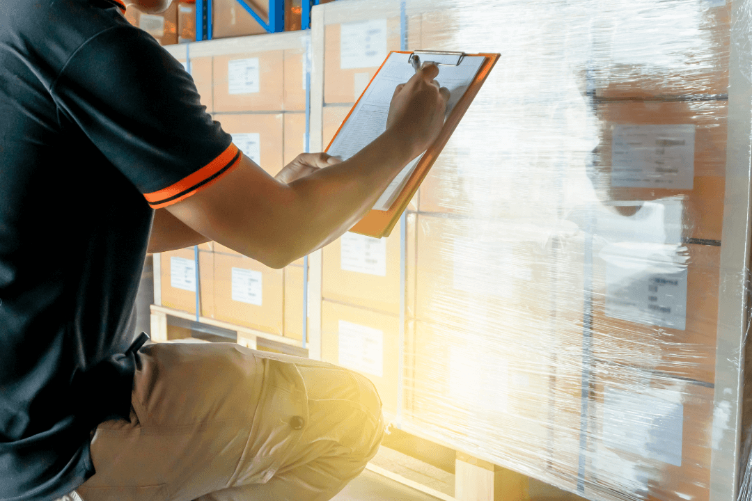 A person in a black shirt kneels and writes on a clipboard while checking a stack of boxes wrapped in plastic in a warehouse.