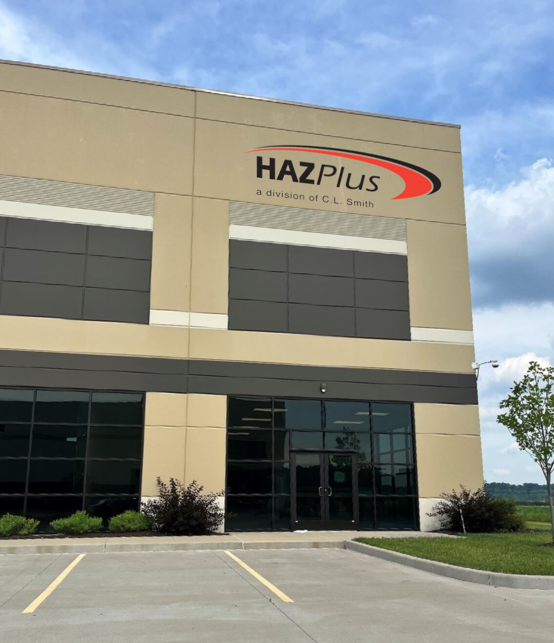 HP New Building MU The exterior of a commercial building with a sign that reads "HAZPlus, a division of C.L. Smith." The entrance features glass doors and windows, with some landscaping and an empty parking lot in front.